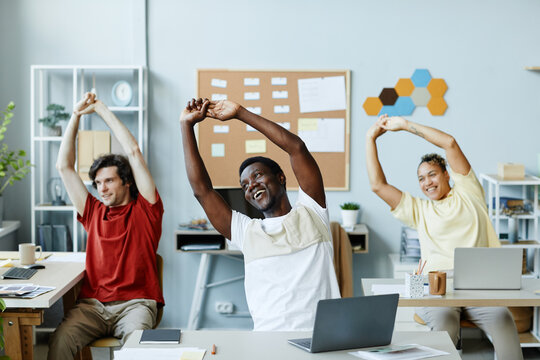 Diverse group of smiling young people doing stretching exercises at workplace in office