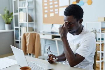 Portrait of frustrated black man taking off glasses at workplace and suffering from headache