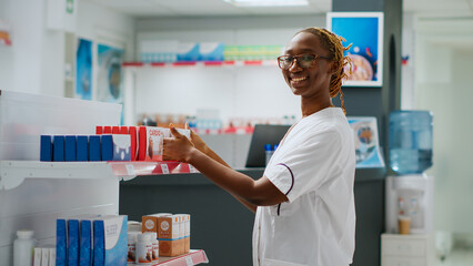 Smiling african american pharmacist checking drugs on shelves in pharmacy, helping clients with...