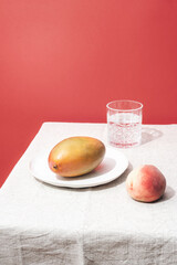 Ripe mango on a plate, peach fruit and a glass of water on a table against red wall. Still life