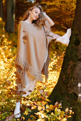 She threw her leg high on a tree in the autumn forest. A beautiful woman in a brown poncho on a background of yellow leaves.