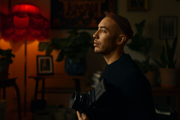 Profile of a man wearing an orange beanie holding a vintage camera at home.