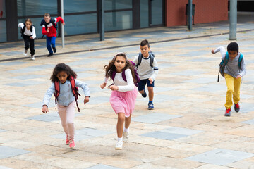 Happy tween schoolchildren of different nationalities with backpacks running together to school lessons outdoors on fall day.