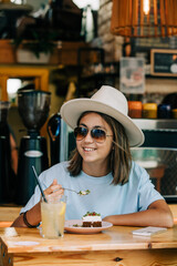 Stylish teenage girl in sunglasses and hat sits in cafe and eats cake