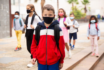 Portrait of schoolboy in medical mask standing near school, kids on background