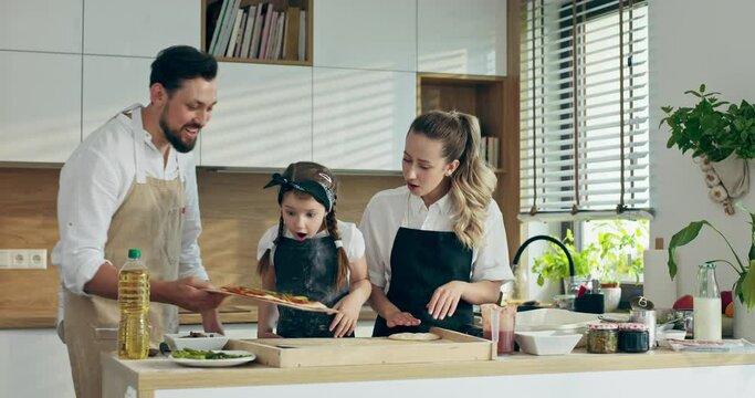 Handsome young father in apron taking homemade freshly baked pizza fron oven putting on wooden desk. Curious delighted mother and daughter watching process waiting for tasting domestic pizza.