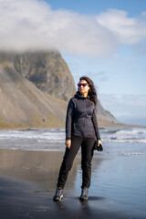 adventurous woman with cap and sunglasses walking on the black volcanic sand beach at the seashore with mountains in autumn winter