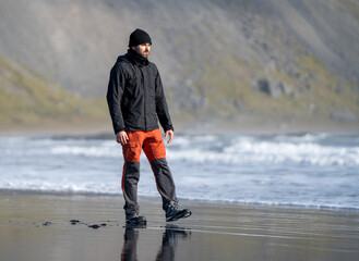 adventurous man with cap and beard walking on a volcanic black sand beach next to mountains in autumn winter