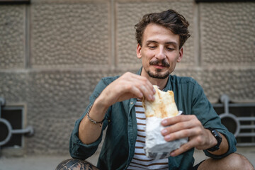 One man young adult modern caucasian male in the city in sunny day stand and eat sandwich fast food...