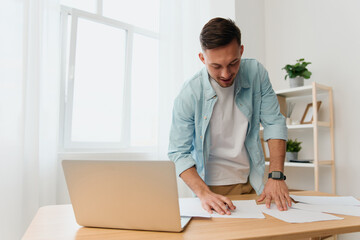 Friendly happy good-looking young businessman reclines on table signs documents looks down smiling in office. Copy space for ad. Remote Job, Technology And Career Profession Concept