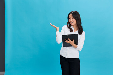Happy cute Asian businesswoman in classic office dress code holds folder tablet point hand up at cope space posing isolated on over blue studio background. Cool business offer. Job interview concept
