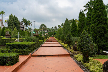 A wide straight stone path with steps among palm trees and trimmed bushes in a city park. Sky with rain clouds.
