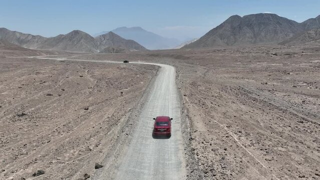 Aerial dolly shot of a red and black vehicle driving along a dusty highway in the desert of peru in front of the holy city of caral with hills, mountains and rocks on a sunny day
