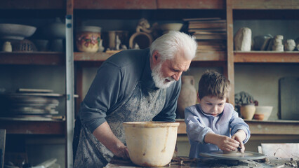 Pottery teacher senior adult in muddy apron is helping young student to form pot from piece of clay...