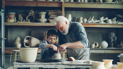 Caring senior grandfather is showing young cute grandson how to work with clay on throwing-wheel in small workshop. Pottery, family hobby and handicraft concept.