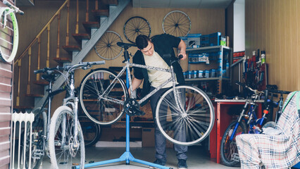 Skilled male mechanic is fixing broken bicycle wheel with wrench then turning it and checking. Many cycles, spare parts, tools and equipment are visible.