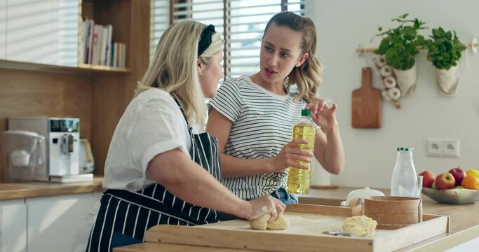 Hapy deligted young daughter helping mother pouring oil on homemade dough while middle aged mother kneading. Women in modern kitchen cooking together having fun while preparing dinner for famly.
