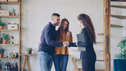 Friendly realtor is giving keys to young couple buyers of new house, happy spouses are hugging , smiling man is shaking hands with broker making deal. - obrazy, fototapety, plakaty