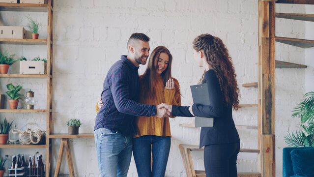 Friendly realtor is giving keys to young couple buyers of new house, happy spouses are hugging , smiling man is shaking hands with broker making deal.