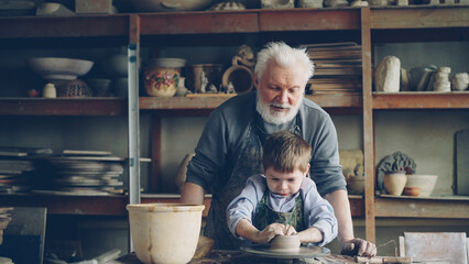 Elderly man experienced potter is teaching cute little boy how to work with clay on potter's wheel....