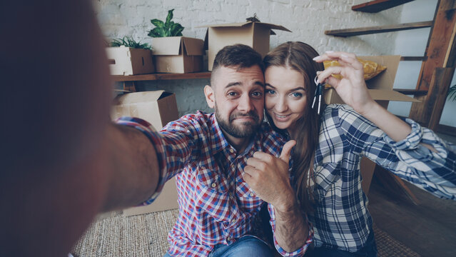 Point of view shot of happy young couple taking selfie with house keys after purchasing new apartment. Young people are holding camera posing and kissing with boxes in background.