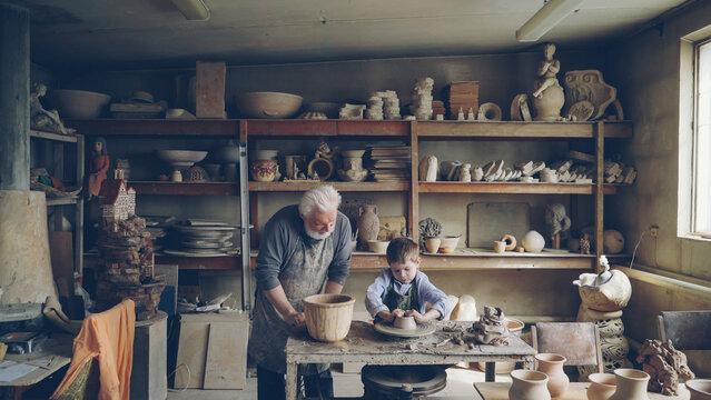 Skilled child is forming pot on potter's wheel while working with his retired grandfather in his figuline home studio. Professional equipment and beautiful ceramic things in background.
