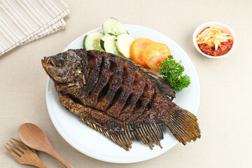 Gurame Bakar Madu, Grilled Gourami with honey and soy sauce. Served with sambal mangga (chilli sauce and grated mango), cucumber and tomatoes in white plate.   
