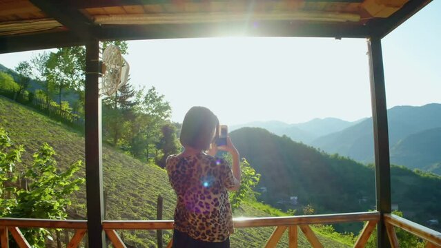 Brunette girl taking pictures of beautiful bright sunset in mountains standing on terrace. Young woman enjoying and admiring beauty of nature and filming amazing landscape. Shooting on the phone