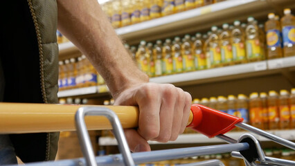 Close-up of a male buyer's hand pushing a supermarket trolley past the rows of oil bottles