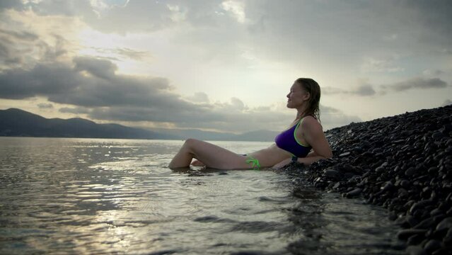 sexy slim young woman in purple swimsuit lies on stone beach near sea or ocean with mountains in background at sunset or dawn. Happy contented girl lies in water and rests on journey.