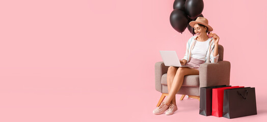 Happy young woman with laptop and shopping bags sitting in armchair on pink background with space...