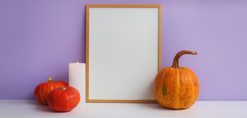 Blank photo frame with Halloween pumpkins and candle on table near lilac wall