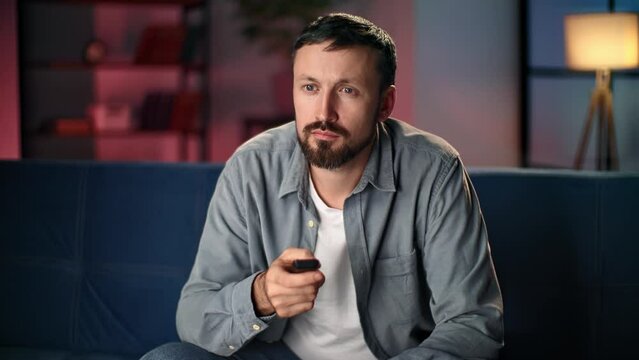 Bored man watching TV at evening home living room checking channels with remote control on couch