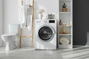 Modern washing machine, toilet bowl and shelf unit with bath accessories near white wall