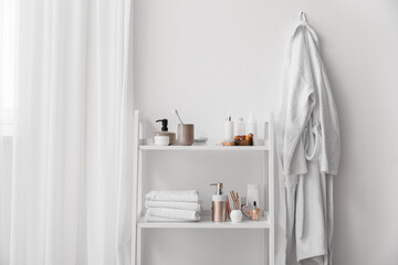 Fototapeta na wymiar Shelf unit with different bath accessories and bathrobe near white wall