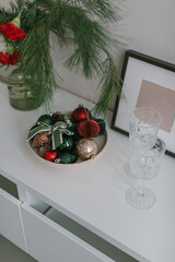 Aesthetic still life with Christmas decor. Christmas toys in ceramic plate,  blank photo frame, glasses on  white dresser. 