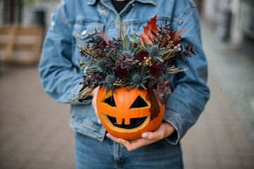 hands of woman holds pumpkin with carved eyes and mouth with flower arrangement inside. Floral decor for halloween.