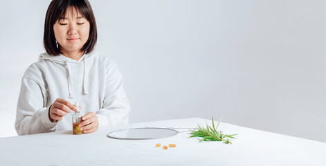 Asian woman with cbd liquid oil made from cannabis extract for a natural skin treatment. Portrait of young woman with cannabis and mirror at the table. Cosmetology and treatment.
