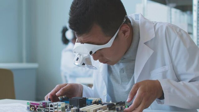 Asian scientist engineer in headband magnifier and lab coat examining motherboard while doing research in laboratory