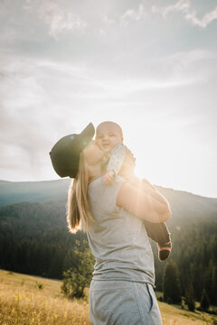 Happy young mother hugs and kisses little baby son standing on autumn grass. Spending time together, outside, on vacation, outdoors. Beautiful sunset in the mountains or in the park.