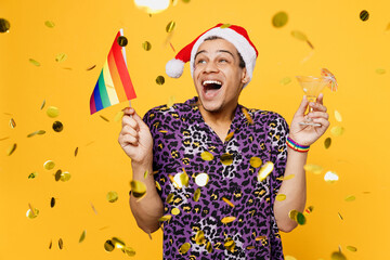 Young happy gay man wear purple animal print shirt Christmas Santa Claus red hat hold martini glass flag among confetti isolated on bright plain yellow color background. Lifestyle lgbtq pride concept.