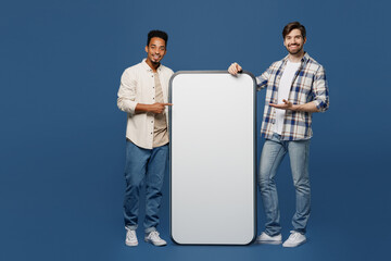 Full body young two friend men wear white casual shirt together show point finger on big huge blank screen mobile cell phone smartphone with mockup area isolated plain dark royal navy blue background