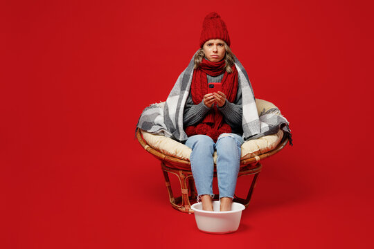 Full size young sad woman wear grey plaid scarf hat sit in chair soar feet use hold mobile cell phone isolated on plain red background Healthy lifestyle ill sick disease treatment cold season concept.