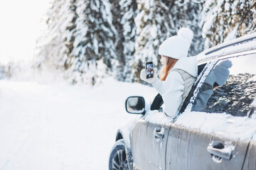 Teenager girl traveling looking out of car window and taking selfie on smartphone in winter snowy forest. Road trip and local travel concept. Happy child enjoying car ride.