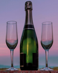 A bottle of champagne with two flute glasses beside it against a beautiful sunset sky