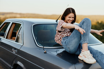 A young woman sits on the trunk of a car, laughs and rests after a difficult road and admires nature with a beautiful view. Stopping is also part of the journey