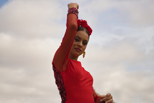 Portrait of young teenage woman in red dance suit with red carnations in her hair doing flamenco poses with clouds in the sky in the background. Flamenco concept, dance, art, typical Spanish dance.