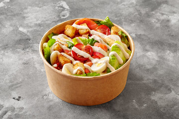 Paper bowl of Caesar salad with lettuce, tomatoes, quail egg, croutons and smoked salmon on gray