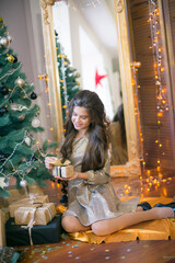 Fototapeta na wymiar A cute teenage girl with long curly hair in a shining dress in a room decorated for Christmas with shining garlands. Christmas mood. beauty and fashion