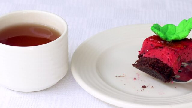 pastry piece slice of cake rectangle shape on white plate and black tea in cup.strawberry cake walnuts orange flower green leaf or pink macaron decoration.fork cutting the desert.tasty morning delight
